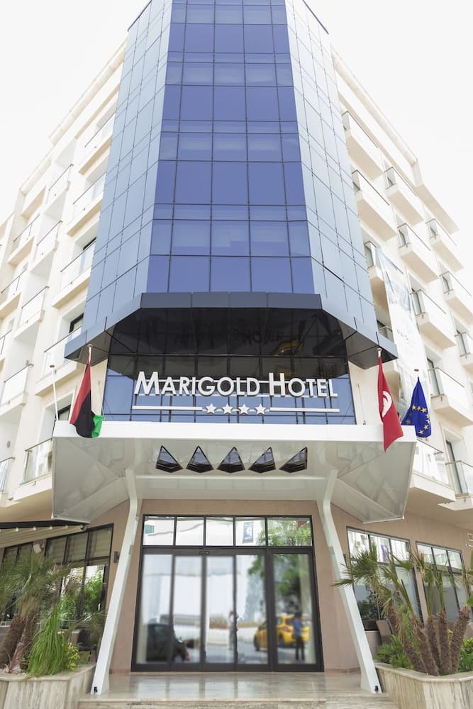 The modern Marigold Hotel entrance, featuring a sleek glass facade and flags, offers a welcoming urban retreat for travelers seeking comfort in a bustling city setting.