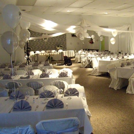 A spacious banquet hall adorned with white balloons and draped ceilings sets a festive mood, featuring neatly arranged tables with blue napkins, ideal for wedding receptions or large gatherings.