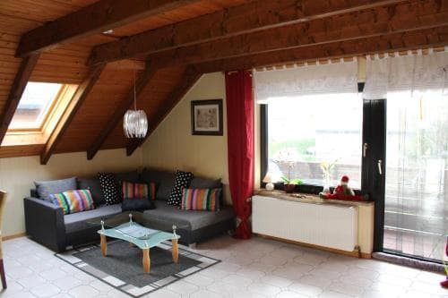 A cozy attic living space with sloped wooden ceilings, colorful cushions on a sofa, a glass coffee table, and a bright window view, ideal for a relaxing vacation rental.