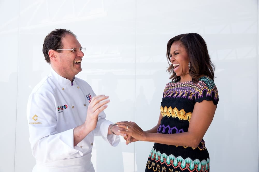 A cheerful interaction unfolds in a bright indoor setting where a chef in a white uniform shares a joyful moment with a woman in a vibrant patterned dress, ideal for cultural exchange events or culinary tourism promotion.