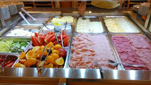 A vibrant buffet spread featuring fresh fruits, sliced meats, and cheeses in a well-lit indoor setting evokes a welcoming dining experience ideal for travelers seeking convenient meal options.