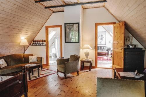 A cozy attic living space with wooden floors, slanted ceilings, and warm lighting creates an inviting atmosphere, featuring comfortable seating, a small table lamp, and a doorway leading to another room, ideal for travelers seeking a charming and intimate accommodation experience.