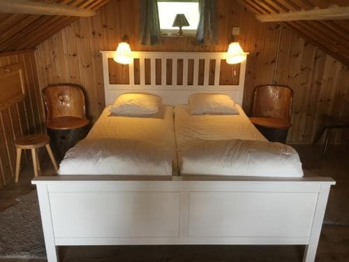A cozy attic bedroom with wooden walls, a neatly made double bed, warm lighting, and rustic furniture evokes a serene retreat ideal for travelers seeking a quaint and intimate accommodation experience.