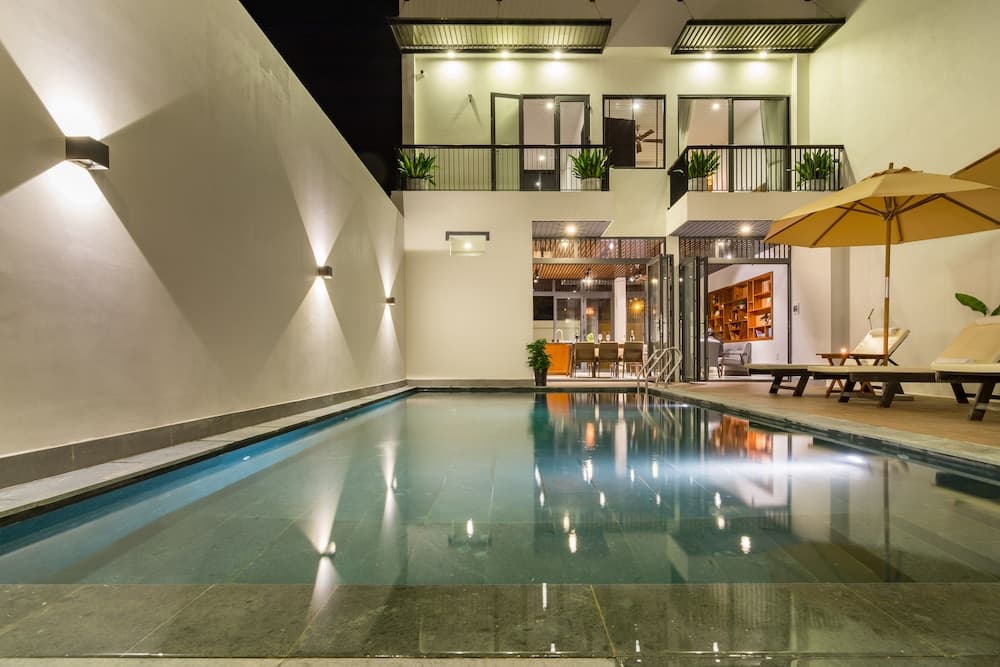 A serene indoor pool area illuminated by soft lighting features lounge chairs under umbrellas, surrounded by modern architecture with balconies and plants, ideal for a luxurious retreat or vacation rental listing.