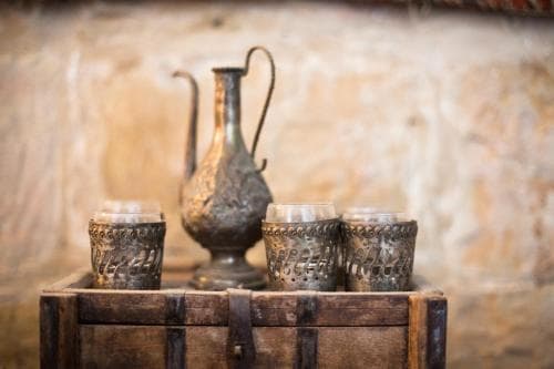 A rustic wooden crate holds ornate metal cups and a traditional pitcher against a textured stone wall, evoking a cozy, historical ambiance ideal for cultural heritage tourism.