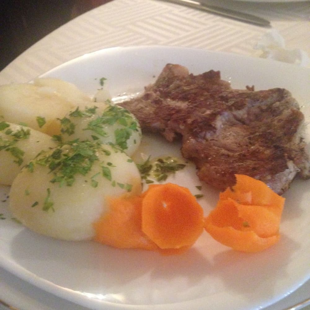 A cozy restaurant plate features a hearty meal of grilled meat, boiled potatoes garnished with parsley, and vibrant carrot slices, perfect for travelers seeking comfort food in a casual dining setting.