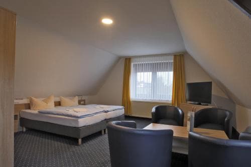 A cozy attic hotel room with a slanted ceiling features a neatly made bed, a small desk with chairs, and a window with yellow curtains, ideal for travelers seeking a quiet and comfortable stay.
