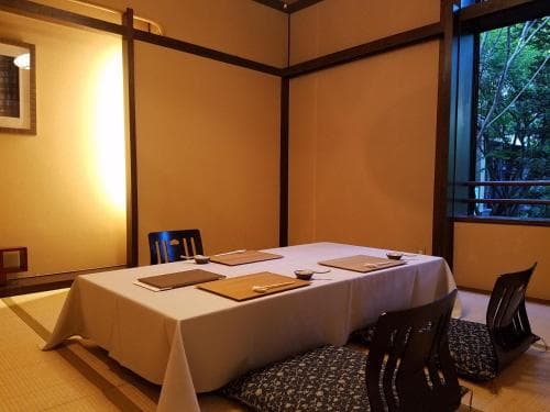 A serene dining room with tatami flooring, a white tablecloth-covered table set for four, and sliding doors revealing lush greenery outside, ideal for travelers seeking a traditional Japanese culinary experience in a tranquil setting.