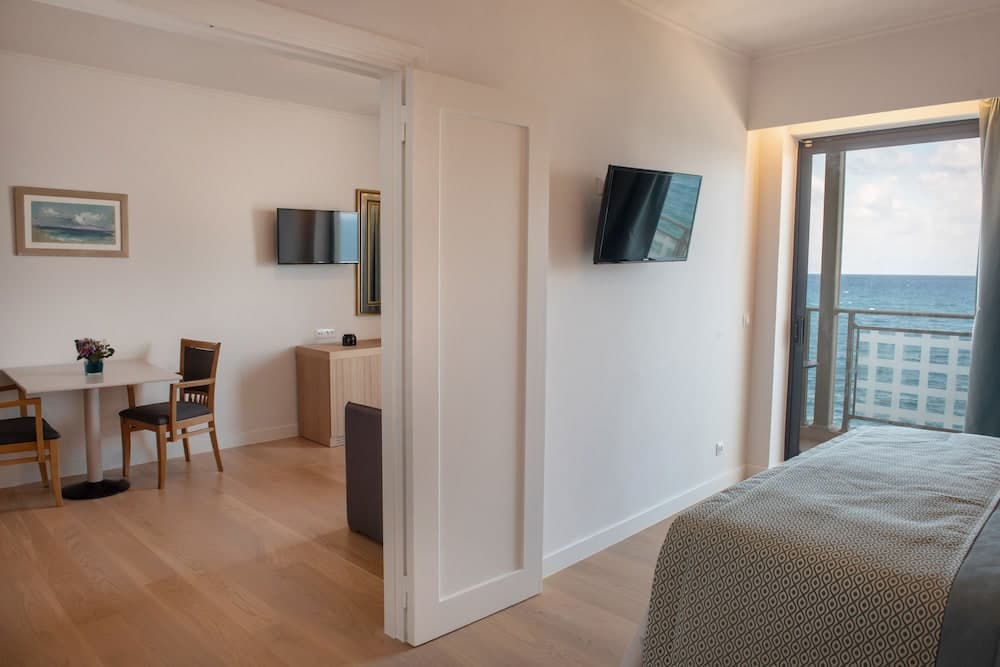 A cozy hotel room with wooden floors, a small dining area, and a balcony overlooking the ocean offers a serene retreat for travelers seeking relaxation and comfort.