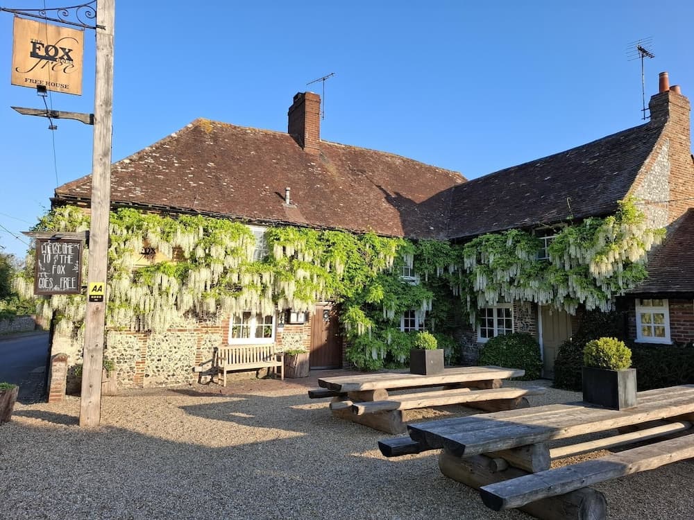 A charming countryside pub adorned with cascading wisteria under a clear blue sky, featuring rustic wooden benches and a welcoming signpost, perfect for travelers seeking a quaint rural retreat.