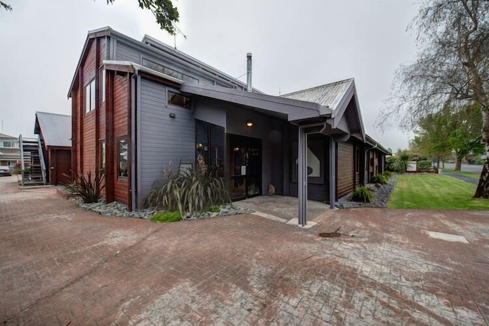A cozy, modern lodge with wooden and metal siding stands invitingly on a paved path amidst greenery, perfect for travelers seeking a serene retreat in a suburban setting.
