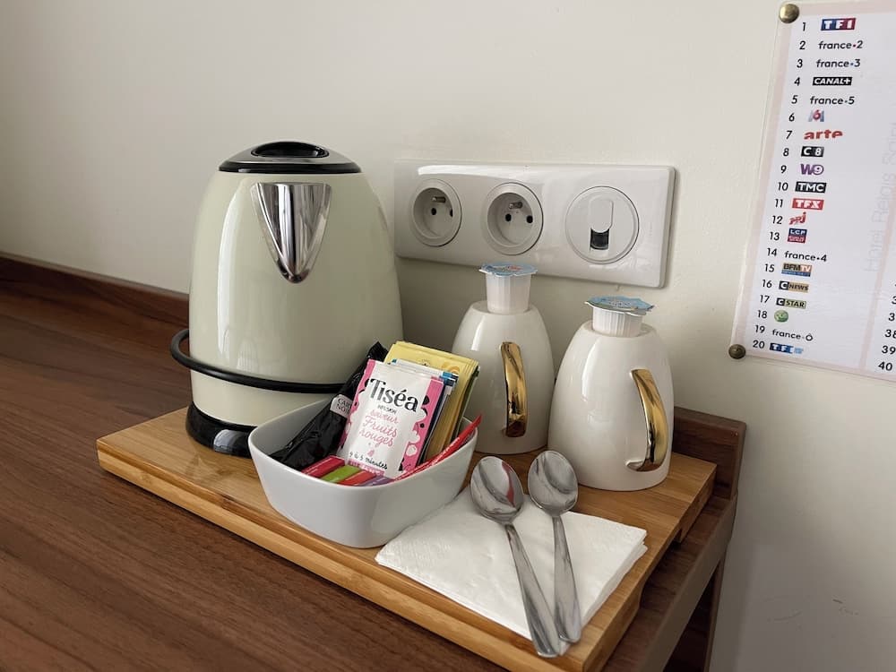 A cozy hotel room corner features a neatly arranged tea station with a kettle, teapots, tea bags, and spoons on a wooden tray, set against a wall with European power outlets and a TV channel guide, ideal for travelers seeking comfort and convenience.