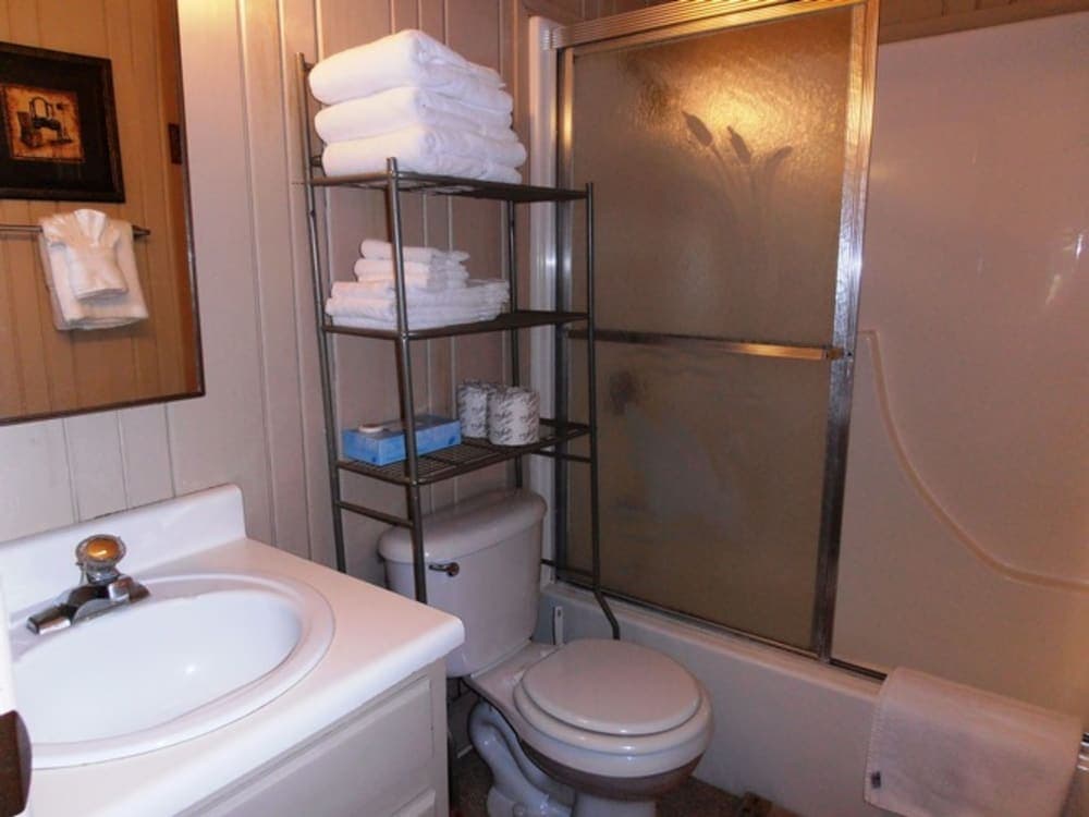 A cozy bathroom with a white sink, toilet, and shower featuring frosted glass doors, complemented by neatly stacked towels on metal shelves, ideal for travelers seeking comfort in compact spaces.