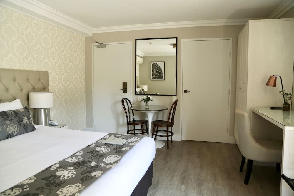 A cozy hotel room features a neatly made bed with floral accents, a small dining area with a round table and chairs, a desk with a lamp, and elegant damask wallpaper, ideal for travelers seeking comfort and functionality.