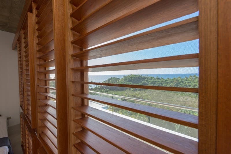 A serene coastal retreat is framed through wooden shutters, offering a tranquil view of lush greenery and the ocean, ideal for travelers seeking relaxation in Las Terrenas, Dominican Republic.