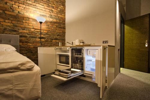 A cozy studio apartment features a rustic brick wall, modern kitchenette with an open dishwasher and fridge, and a comfortable bed, ideal for travelers seeking a compact yet stylish urban retreat.