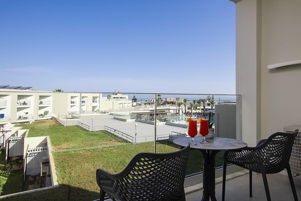 A serene balcony view of a modern resort complex with lush greenery, palm trees, and a pool area under a clear blue sky, featuring two vibrant cocktails on a table, perfect for a relaxing vacation getaway.