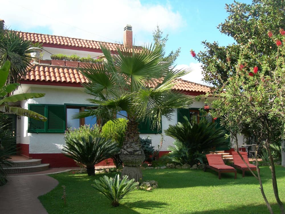 A serene Mediterranean villa garden features lush greenery, palm trees, red lounge chairs, and a tiled-roof house under a bright blue sky, ideal for a relaxing vacation retreat.