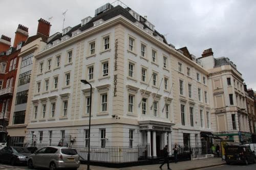 A bustling urban street showcases a grand white hotel with classic architecture, surrounded by pedestrians and vehicles under an overcast sky, ideal for city exploration and accommodation booking.