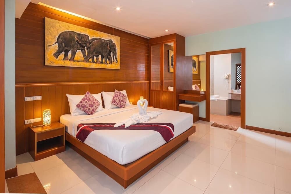 A cozy hotel bedroom features a neatly made bed adorned with swan towels, warm wooden accents, an elephant-themed artwork, and a connected bathroom, ideal for travelers seeking comfort and cultural ambiance.
