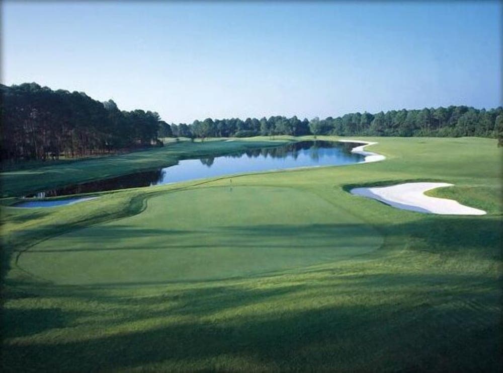 A serene golf course features lush green fairways, a reflective water hazard, and sand bunkers under a clear blue sky, ideal for leisurely golfing getaways.