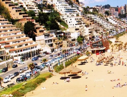 A vibrant coastal cityscape unfolds with sandy beaches dotted by straw umbrellas, bustling streets lined with parked cars, and densely packed hillside buildings under a sunny sky, ideal for beachgoers seeking relaxation and urban exploration.