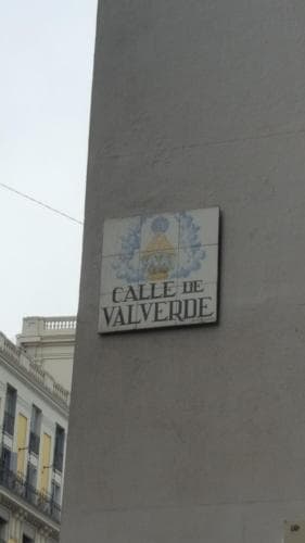 A serene urban scene captures the "Calle de Valverde" street sign on a pillar, set against classic European architecture, ideal for travelers exploring historic cityscapes.