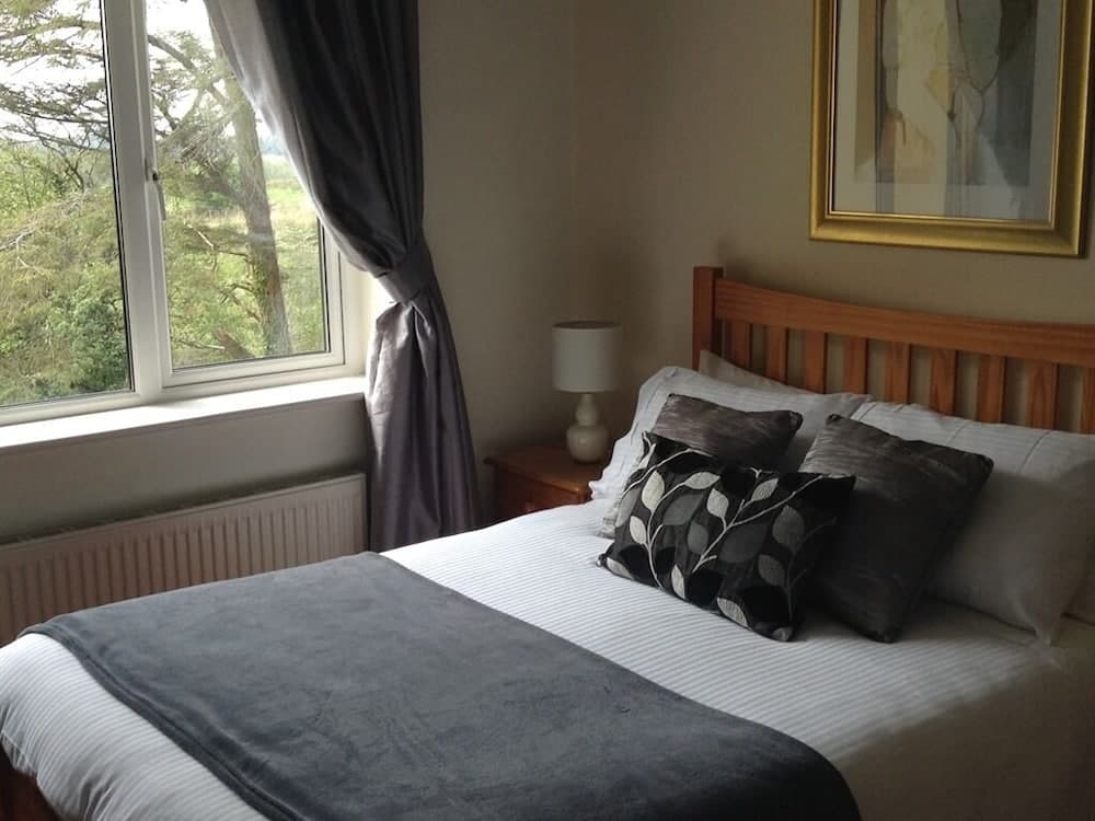 A cozy bedroom with a neatly made bed adorned with patterned pillows, a wooden headboard, and a window offering a serene view of greenery, ideal for travelers seeking relaxation in a tranquil setting.