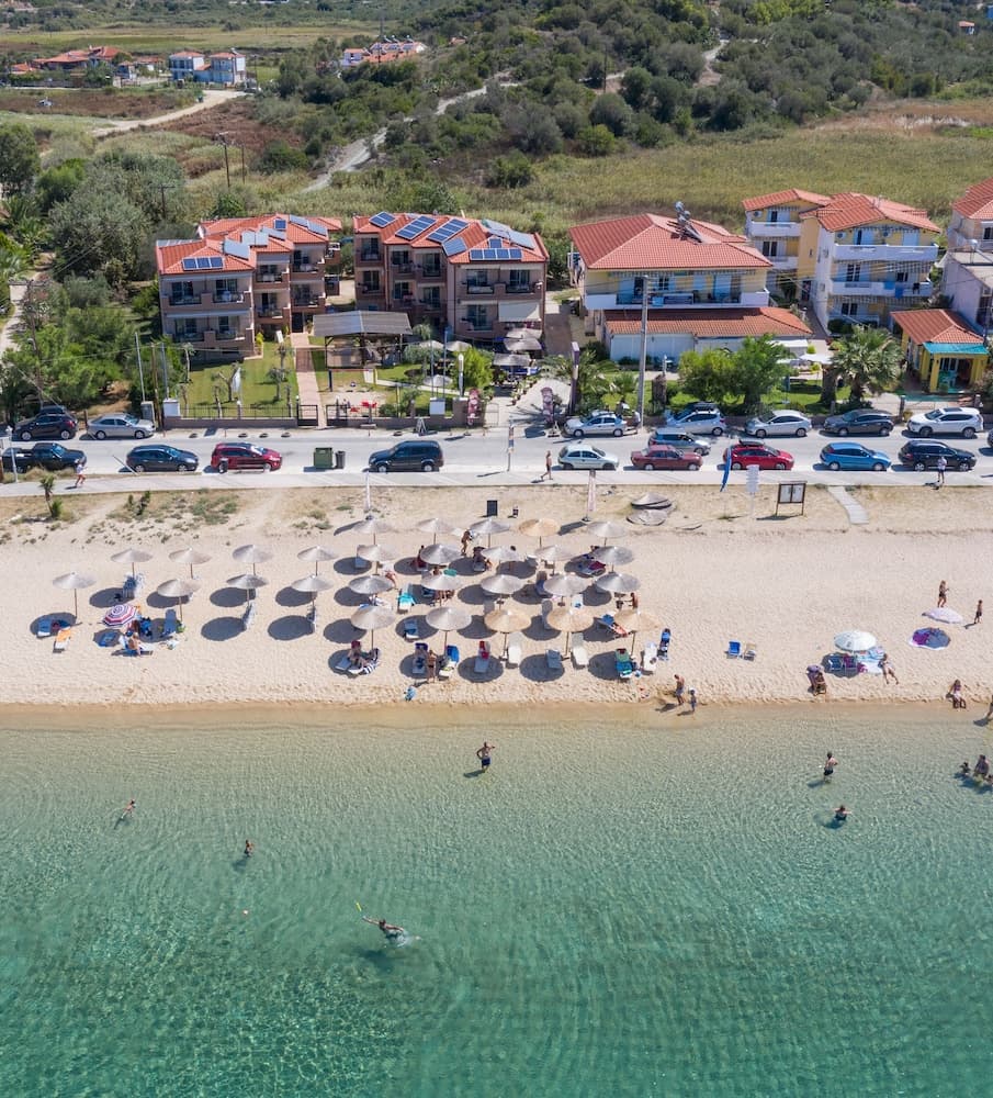 A vibrant coastal beach scene features clear turquoise waters, sunbathers under straw umbrellas, and colorful buildings in the background, ideal for a relaxing seaside getaway.