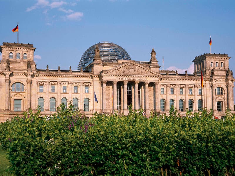 A majestic historical building with a glass dome and German flags stands under a clear blue sky, surrounded by lush greenery, ideal for cultural exploration and sightseeing tours.