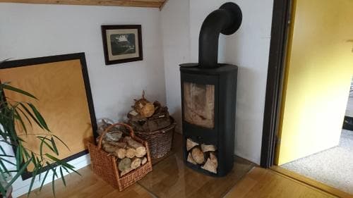A cozy indoor scene featuring a wood-burning stove surrounded by neatly stacked firewood in baskets, set against a warm-toned wall with framed artwork, ideal for travelers seeking rustic charm and comfort in a cabin or countryside retreat.