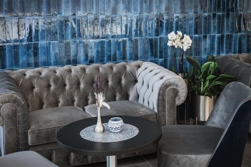 A cozy lounge area features plush gray velvet sofas against a striking blue textured wall, adorned with elegant floral arrangements and a small table, ideal for travelers seeking a sophisticated relaxation spot in a boutique hotel.