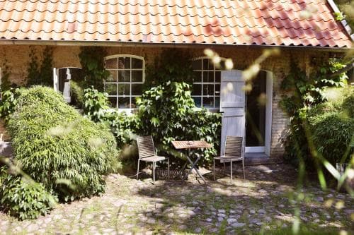 A charming rustic cottage with a red-tiled roof and ivy-covered walls offers a serene outdoor seating area on a cobblestone patio, ideal for a peaceful countryside getaway.