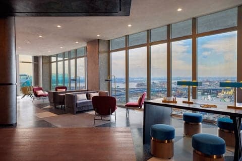 A modern hotel lounge with panoramic city views through large windows, featuring stylish seating and vibrant decor, ideal for travelers seeking relaxation and urban ambiance in Flushing, NY.