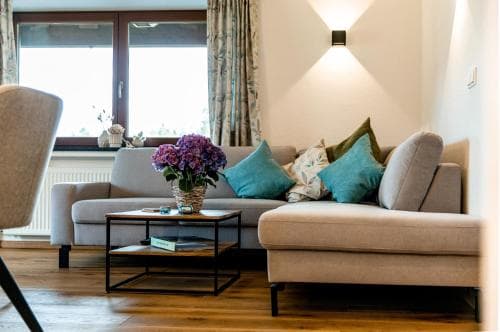 A cozy living room with a plush sofa adorned with teal cushions, a vibrant floral centerpiece on a modern coffee table, and large windows draped with patterned curtains, evokes a serene and inviting atmosphere ideal for travelers seeking comfort in a vacation rental.
