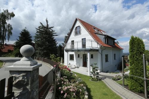 A charming white house with a red-tiled roof stands amidst lush greenery under a partly cloudy sky, offering a serene suburban retreat ideal for family vacations.