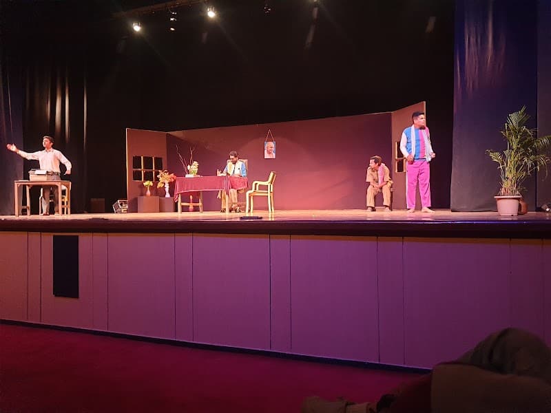 A vibrant theatrical performance unfolds on the stage of Kamani Auditorium in New Delhi, featuring actors engaged in a lively scene with props like tables, chairs, and potted plants, ideal for cultural enthusiasts exploring local arts and entertainment.