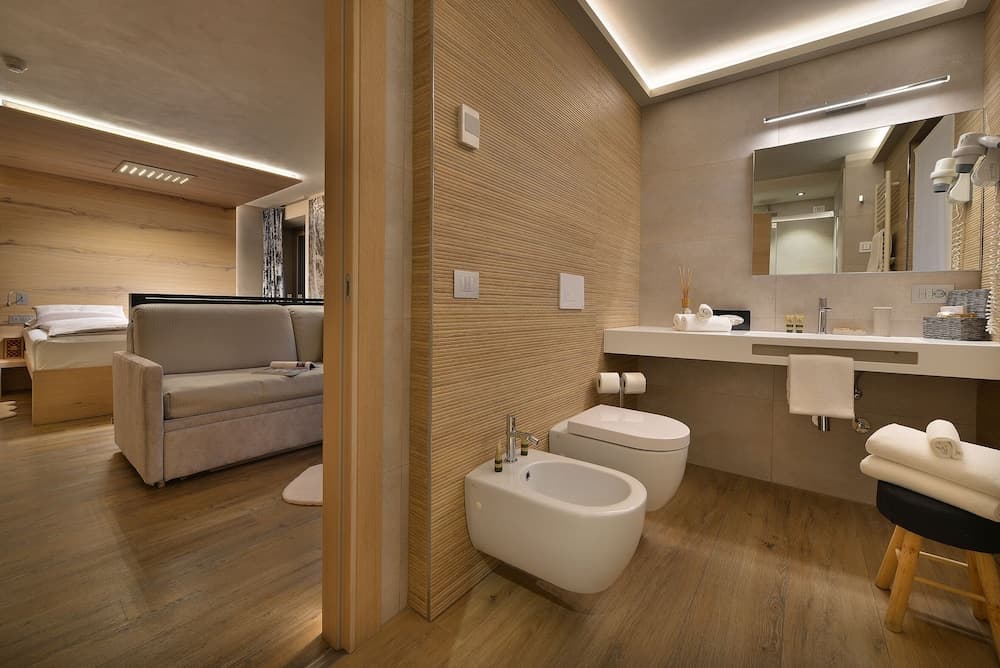 A cozy hotel suite features a modern bathroom with a bidet, sleek vanity, and warm wooden accents, creating an inviting atmosphere ideal for travelers seeking comfort and style.