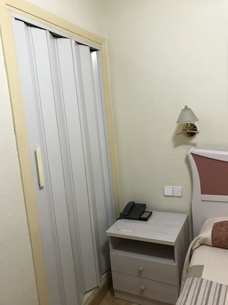 A cozy hotel room corner features a neatly arranged bedside table with a phone and lamp, a closed closet door, and soft lighting, ideal for travelers seeking comfort and convenience.