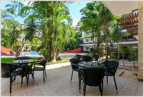 A serene tropical resort patio features black wicker tables and chairs under lush palm trees, with a sparkling pool and modern buildings in the background, ideal for travelers seeking relaxation and leisure.