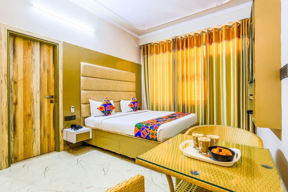 A cozy hotel room features a neatly made bed adorned with colorful pillows, golden curtains, a wicker dining set with refreshments, and warm lighting, ideal for travelers seeking comfort and relaxation.