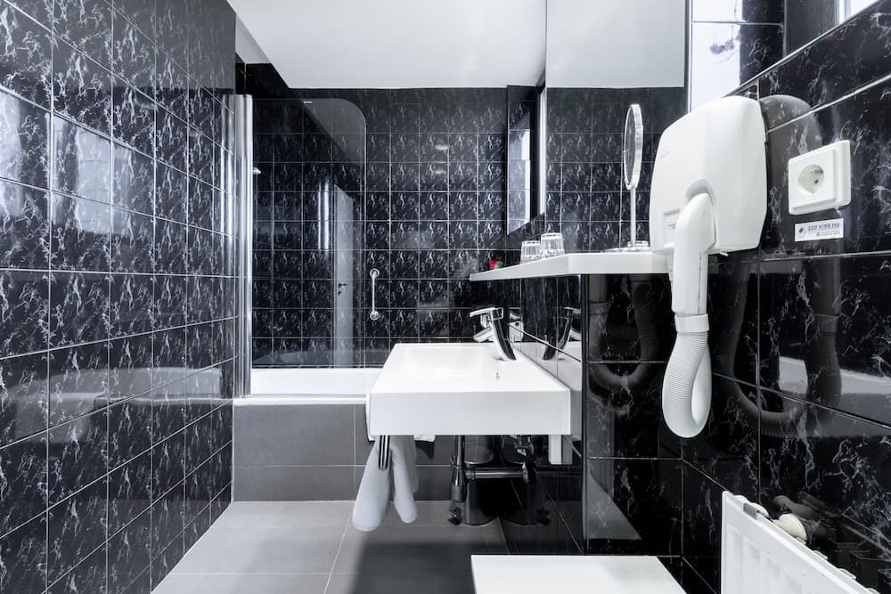 A sleek, modern bathroom with black marble tiles exudes elegance, featuring a white sink, wall-mounted hairdryer, and glass shower enclosure, ideal for travelers seeking luxury accommodations.