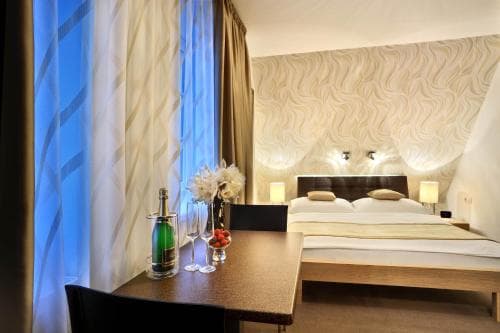 A cozy hotel room features a neatly made bed with patterned headboard lighting, a small table adorned with flowers and a champagne bottle, set against soft curtains, ideal for a romantic getaway or business trip.