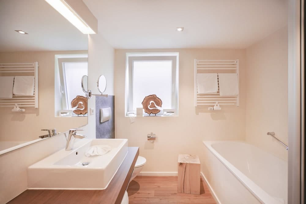 A serene hotel bathroom features a modern sink, wooden floor, bathtub, towel racks, and a heart-shaped wooden decoration by the window, ideal for travelers seeking comfort and relaxation.