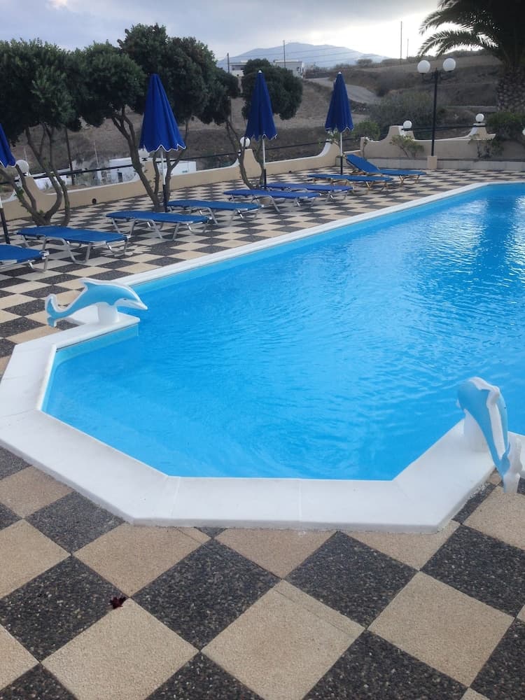 A serene resort pool area features a checkered patio, blue lounge chairs, closed umbrellas, dolphin sculptures, and lush greenery, ideal for relaxing vacations.