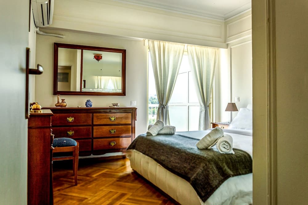 A cozy hotel bedroom with warm wooden floors, a neatly made bed adorned with folded towels, a vintage dresser topped with decorative items, and soft natural light filtering through sheer curtains, ideal for travelers seeking comfort and charm.