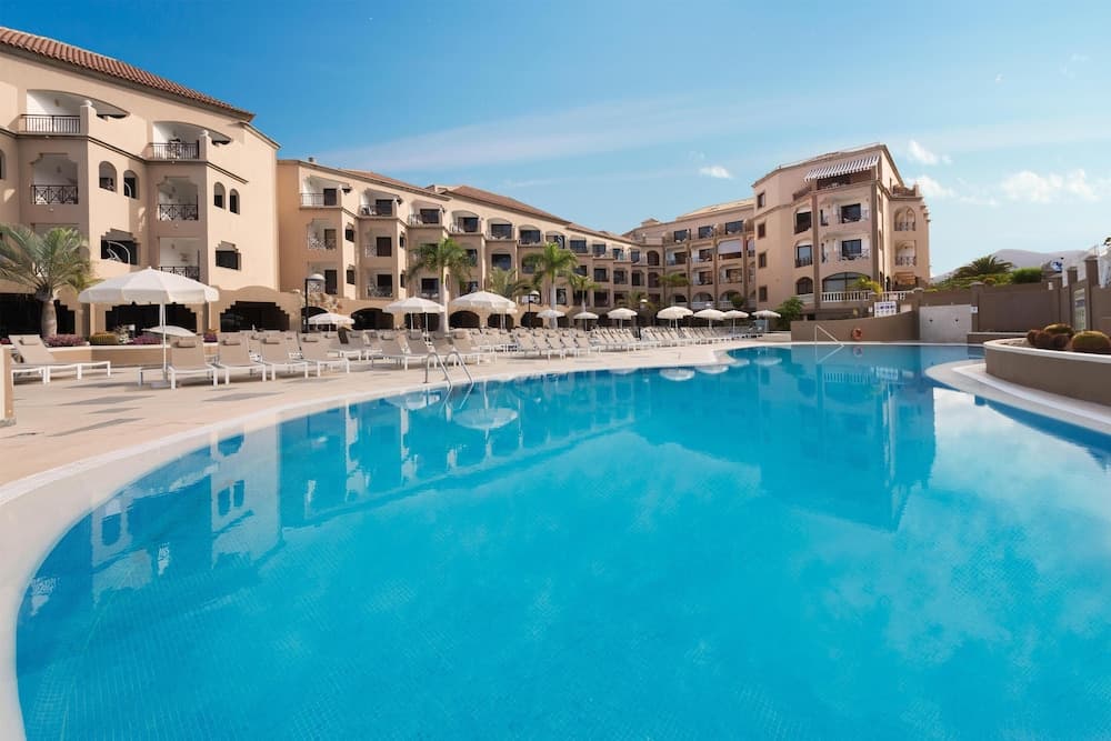 A serene resort scene features a sparkling blue pool surrounded by lounge chairs and umbrellas, set against multi-story buildings under a clear sky, ideal for vacationers seeking relaxation and leisure.