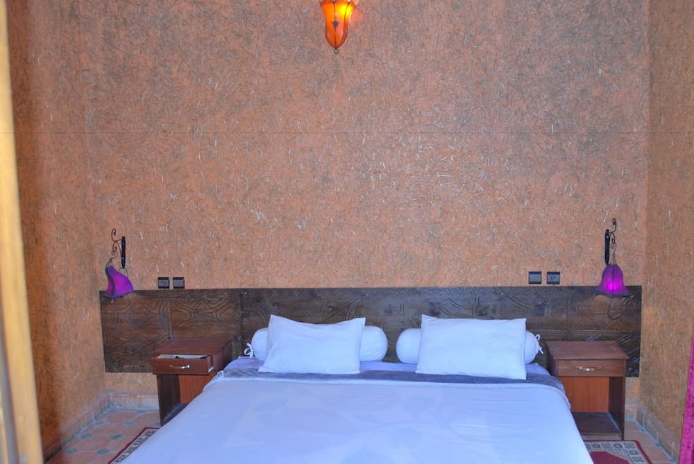 A cozy hotel room features a neatly made bed with white linens against a textured orange wall, illuminated by warm purple lamps, ideal for travelers seeking comfort and relaxation.
