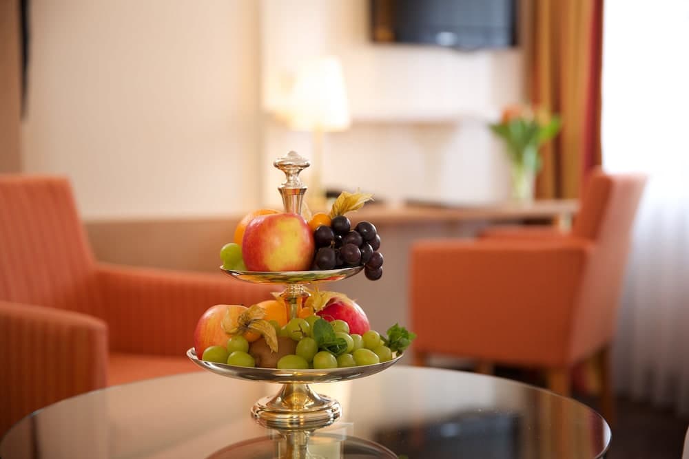 A cozy hotel lounge features a vibrant two-tiered fruit display on a glass table, surrounded by orange armchairs and soft lighting, creating an inviting atmosphere ideal for relaxation or business meetings.
