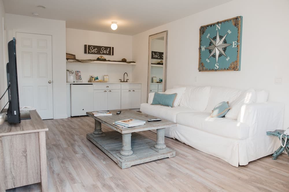 A cozy beach-themed living room with white furniture, wooden floors, nautical decor including a compass wall art and "Set Sail" sign, ideal for a relaxing coastal vacation rental.